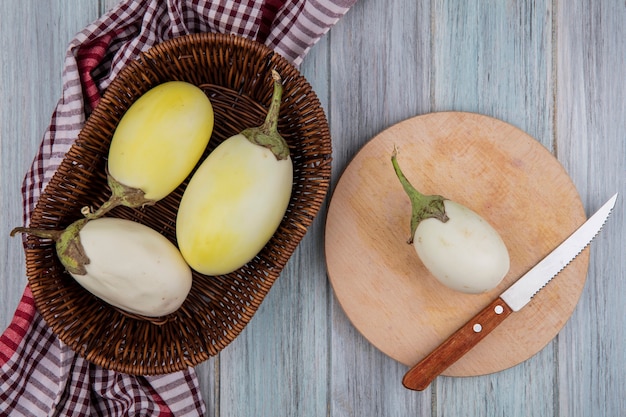 Top view of yellow and white eggplants with knife on cutting board and in basket on plaid cloth on wooden background
