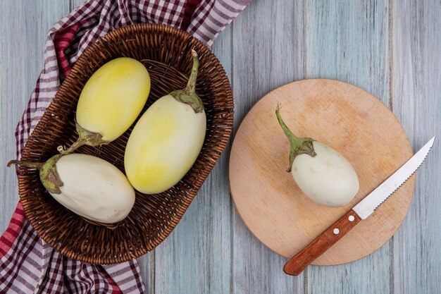 Top view of yellow and white eggplants with knife on cutting board and in basket on plaid cloth on wooden background