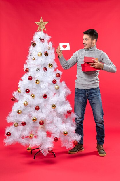 Top view of young adult in a gray blouse standing near the decorated white Christmas tree and holding his gifts on red