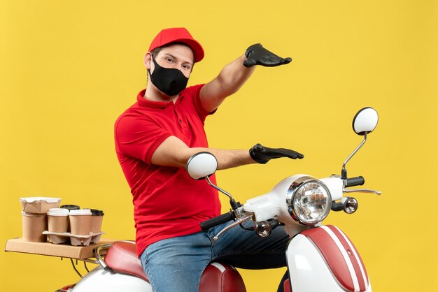 Top view of young adult wearing red blouse and hat gloves in medical mask delivering order sitting on scooter feeling confident on yellow background