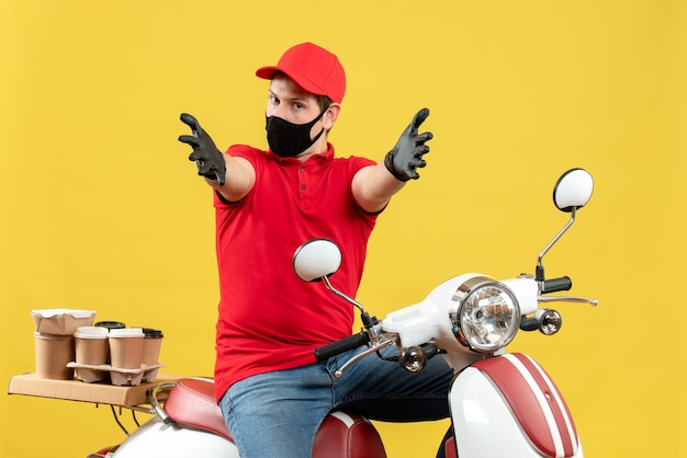 Вид сверху молодого человека в красной блузке и шляпных перчатках в медицинской маске, доставляющего заказ, сидя на скутере, чувствуя замешательство, протягивая руки вперед на желтом фоне