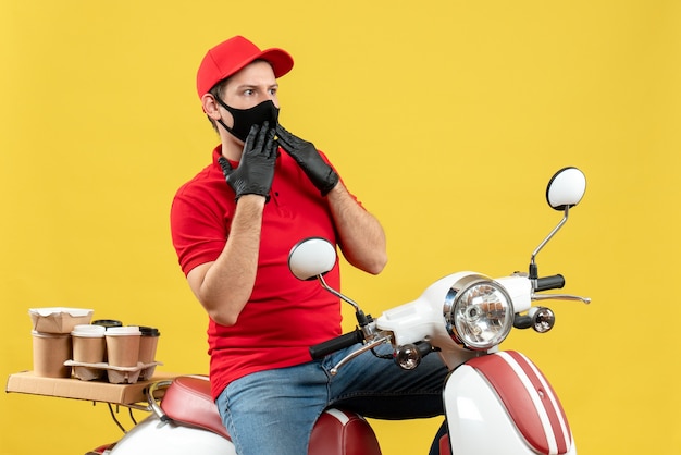 Top view of young adult wearing red blouse and hat gloves in medical mask delivering order sitting on scooter focused on something on yellow background