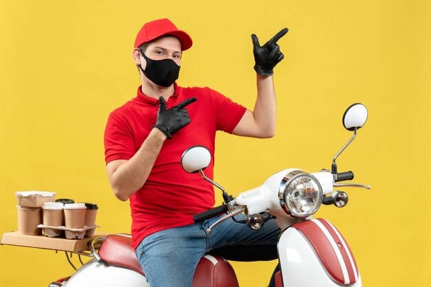 Top view of young adult wearing red blouse and hat gloves in medical mask delivering order sitting on scooter pointing up on yellow background