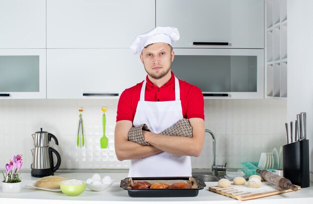 Top view of young chef wearing holder standing behind the table with pastries eggs grater on it in the white kitchen