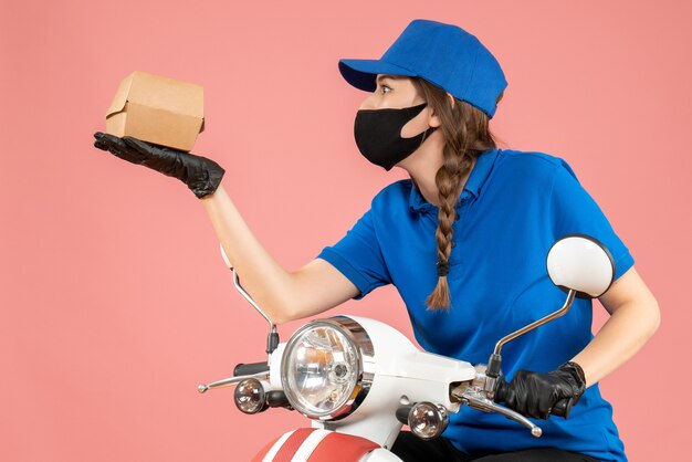 Top view of young concentrated female courier wearing medical mask and gloves holding box on pastel peach