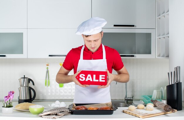 Top view of young concentrated male chef showing sale sign in the white kitchen