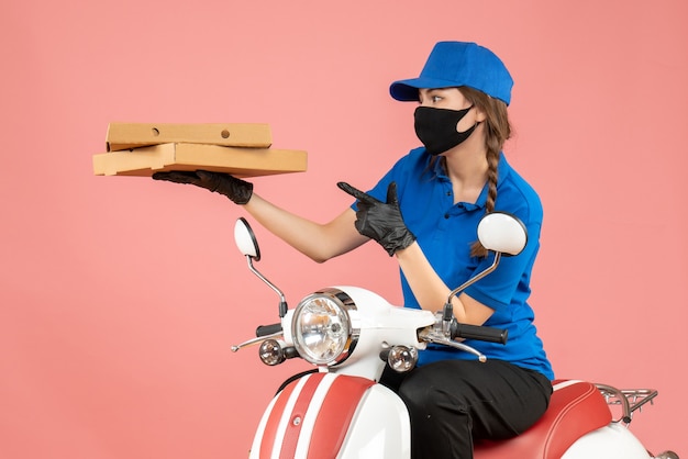Top view of young confused female courier wearing medical mask and gloves sitting on scooter delivering orders on pastel peach