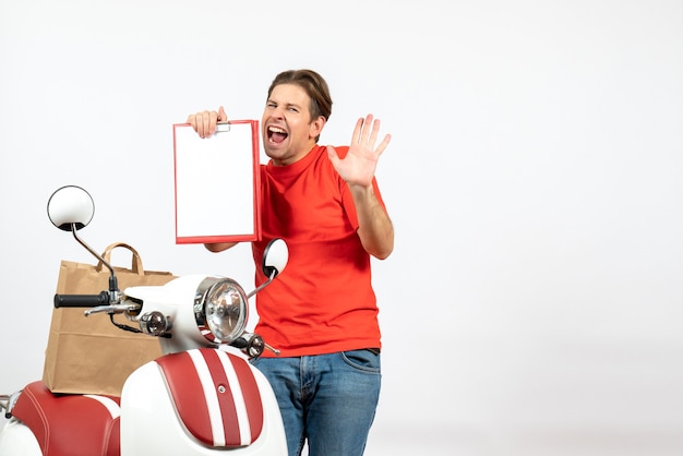 Top view of young emotional nervous delivery guy in red uniform standing near scooter showing document on white wall