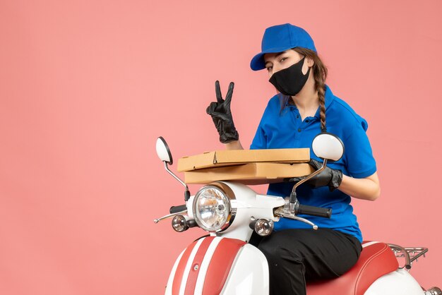 Top view of young female courier wearing medical mask and gloves sitting on scooter delivering orders showing two on pastel peach