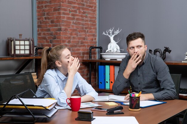 Top view of young female worker and her male co-worker sitting at the table yawning in office enviroment