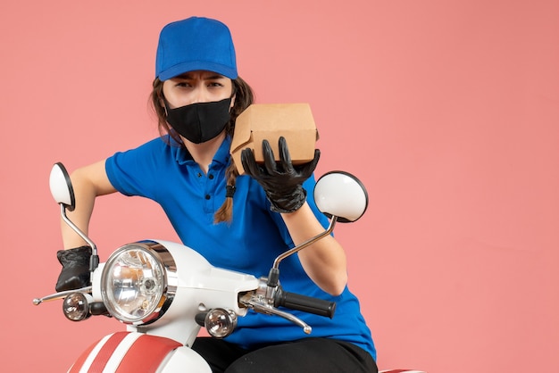 Top view of young focused female courier wearing medical mask and gloves holding box on pastel peach