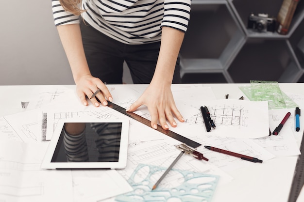 Free photo top view of young good-looking architect student girl in casual striped shirt and black jeans standing near table, holding ruler and pen in hands making drawings, watching movie on digital table, gett