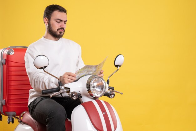 Top view of young guy sitting on motorcycle with suitcase on it looking at map on isolated yellow background