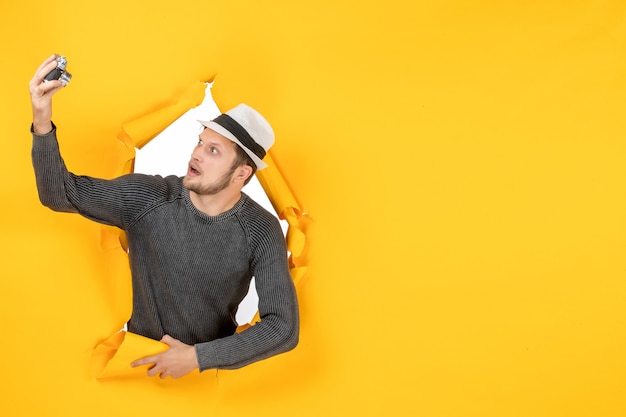 黄色い壁に引き裂かれたカメラを押しながら見ている帽子をかぶった若い男のトップビュー
