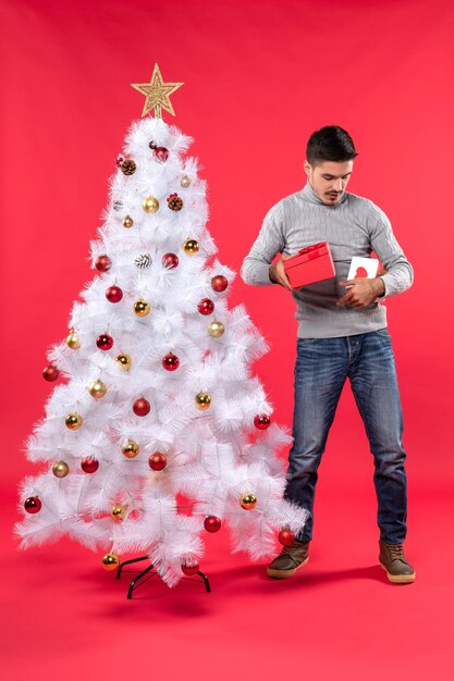 Top view of young handsome adult in a gray blouse standing near the decorated white Christmas tree