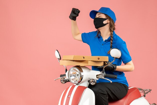 Top view of young happy emotional female courier wearing medical mask and gloves sitting on scooter delivering orders on pastel peach
