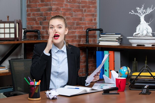 Top view of young lady sitting at a table and holding the document calling someone in the office