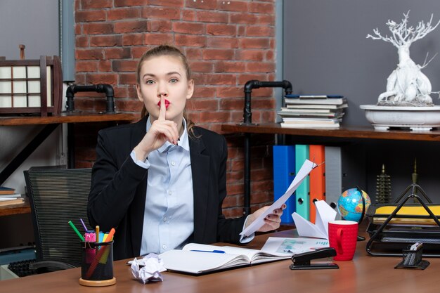 Top view of young lady sitting at a table and holding the document making silence gesture in the office