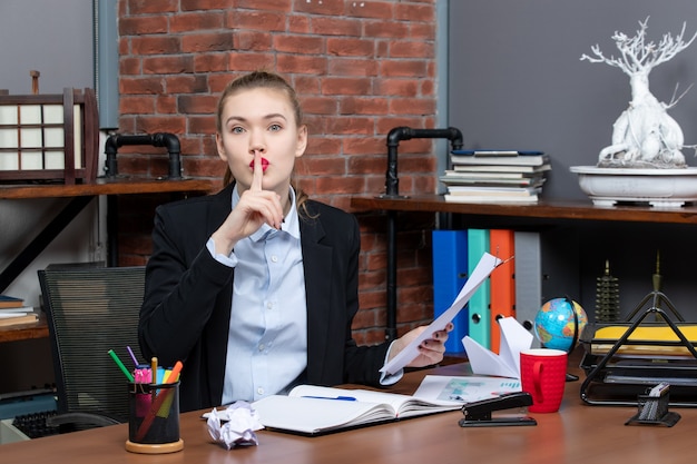 Top view of young lady sitting at a table and holding the document making silence gesture in the office