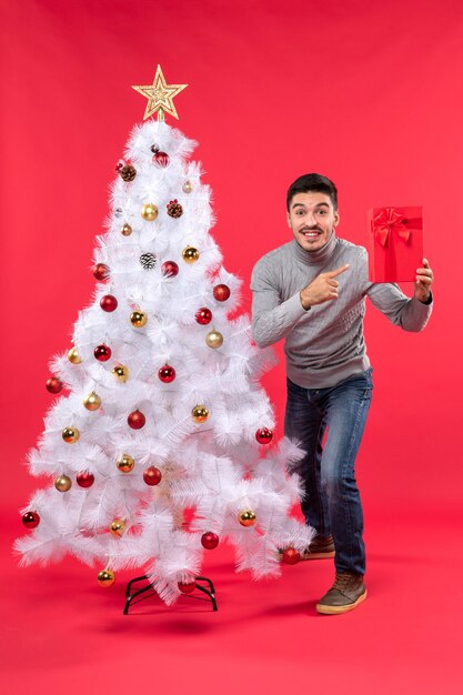 Top view of young man standing near the decorated white New Year tree and holding his gifts