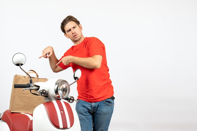 Top view of young shocked delivery guy in red uniform standing near scooter showing document on white wall