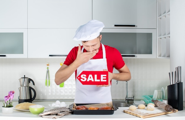 Foto gratuita vista dall'alto del giovane chef maschio assonnato che mostra il cartello di vendita nella cucina bianca white