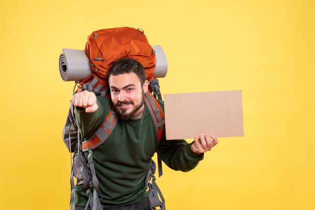 Top view of young smiling and confident travelling guy with backpack holding a sheet without writing on yellow