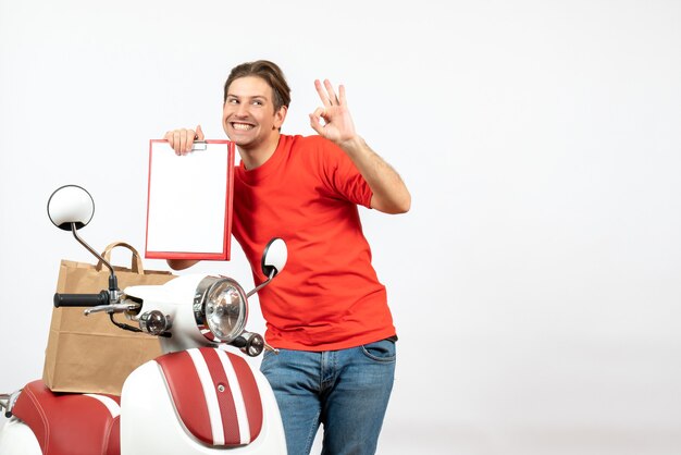 Top view of young smiling delivery guy in red uniform standing near scooter showing document making eyeglasses gesture on white wall