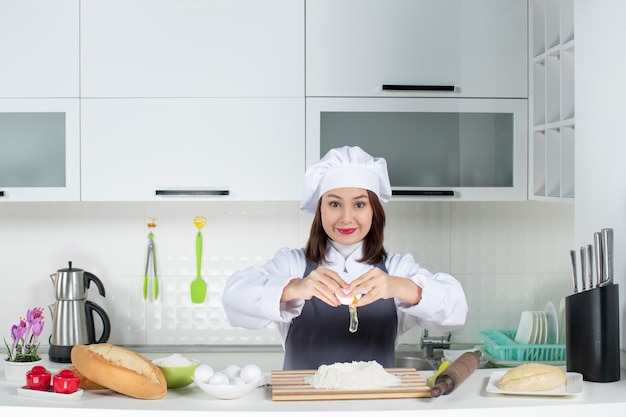 白いキッチンで卵を食べ物に割ってテーブルの後ろに立っている制服を着た若い笑顔の女性シェフの上面図