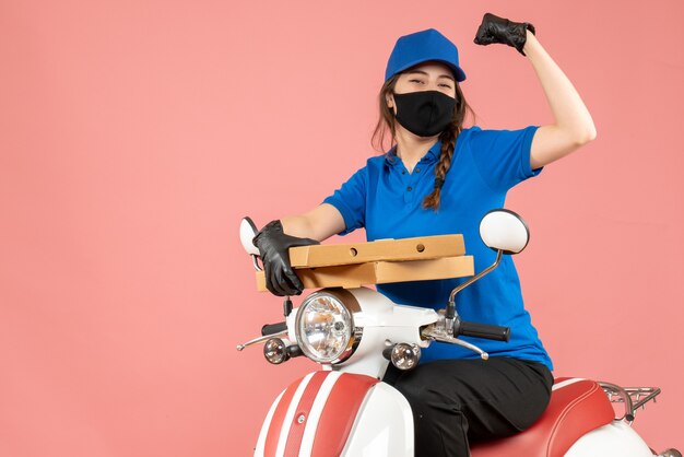 Top view of young smiling happy female courier wearing medical mask and gloves sitting on scooter delivering orders on pastel peach