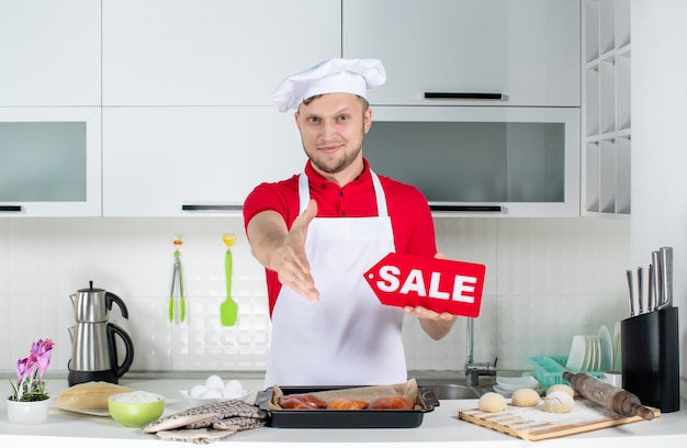 Vista dall'alto di un giovane chef maschio sorridente che tiene in mano un cartello di vendita e dà il benvenuto a qualcuno nella cucina bianca