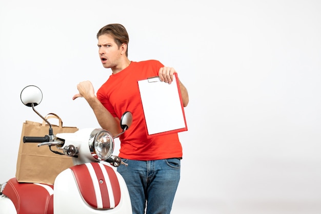 Top view of young surprised delivery guy in red uniform standing near scooter showing document pointing back on white wall