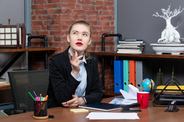 Вид сверху молодой вдумчивой женщины-офисного работника, сидящей за своим столом и позирующей перед камерой