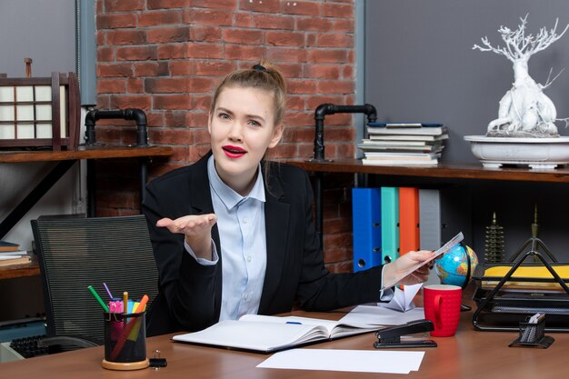 Top view of young woman sitting at a table and holding a document curious about something in the office