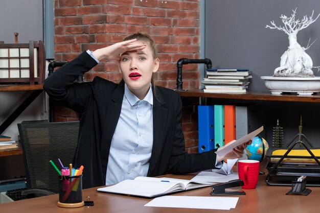 Top view of young woman sitting at a table and holding a document looking at something carefully in the office