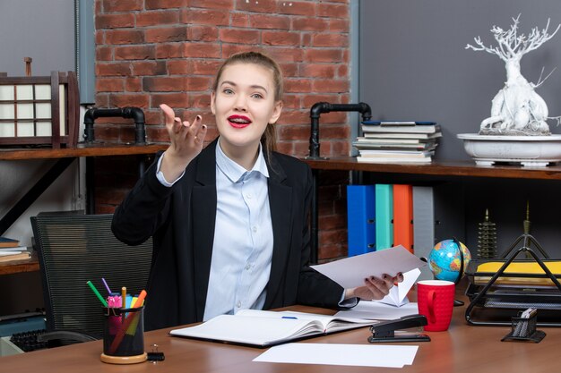 Top view of young woman sitting at a table and holding a document looking at something carefully with smiling facia expression in the office