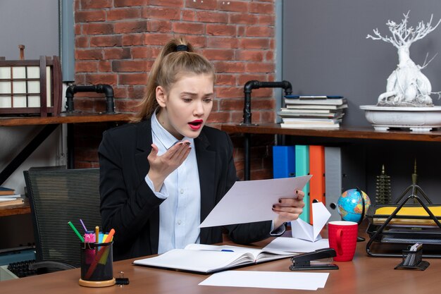 Top view of young woman sitting at a table and holding a document reading something with confused facial expression in the office