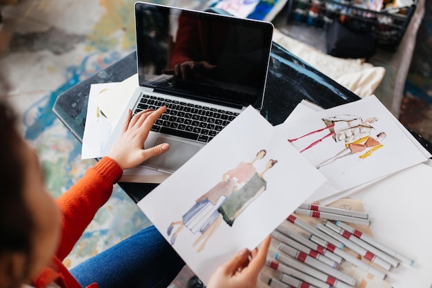 Top view of young woman sitting at the table working on laptop with fashion illustrations near in modern workshop