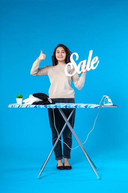 Вид сверху молодой женщины, стоящей за гладильной доской и показывающей значок продажи, делая жест "ОК" на синем фоне