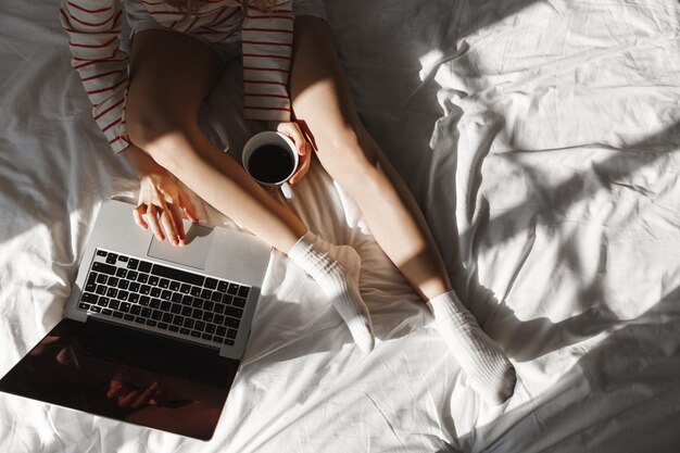 Top view of young woman using laptop in bed drinking coffee and resting at home Cropped shot of female legs in cozy socks girl relaxing on weekends