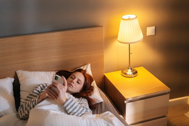 Top view of young woman using smartphone looking on screen typing message lying on bed late at night, bedside lamp lighting with warm yellow light. tired insomnia lady browsing smartphone in bed.