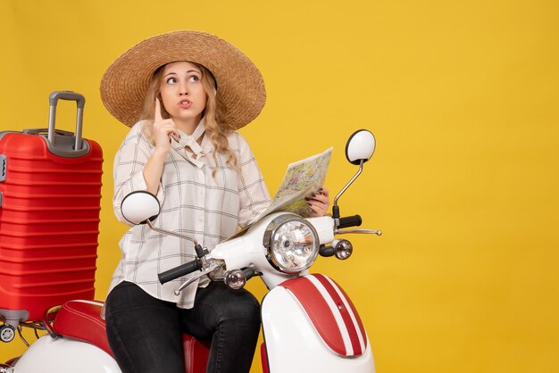 Top view of young woman wearing hat and sitting on motorcycle and holding map pointing up on yellow 