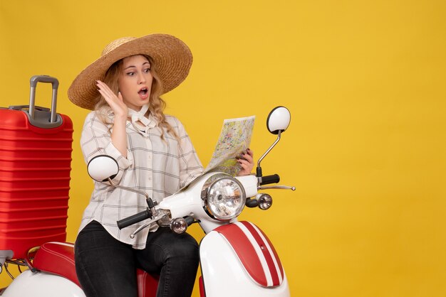 Top view of young woman wearing hat and sitting on motorcycle and looking at map with surprising facial epression on yellow 