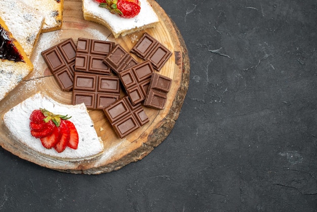 Top view yummy cake slices with strawberries and choco bars on dark surface