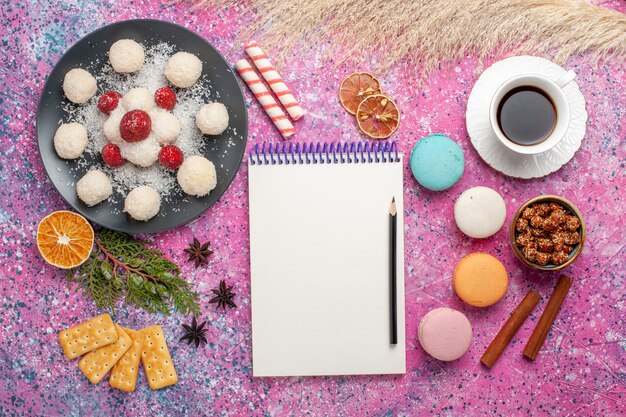 Top view of yummy coconut candies with macarons and cup of tea on light-pink surface