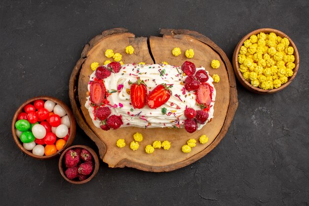 Top view of yummy cream cake with fruits and candies on black