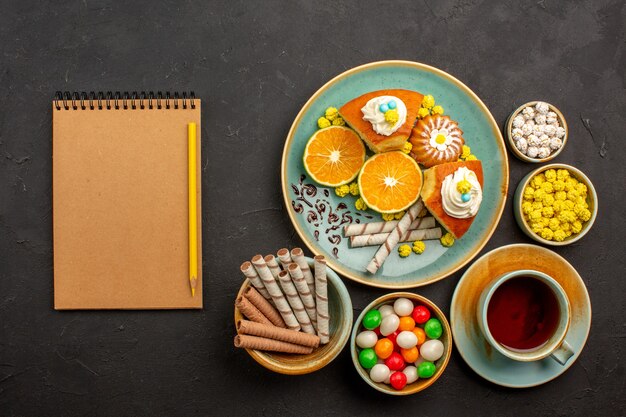 Top view yummy pie slices with fresh tangerines tea and candies on a dark