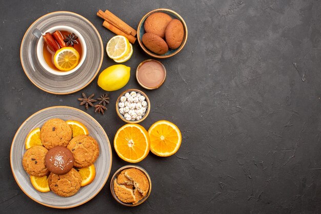 Top view yummy sand cookies with fresh oranges and cup of tea on dark background fruit biscuit sweet cookies citrus sugar