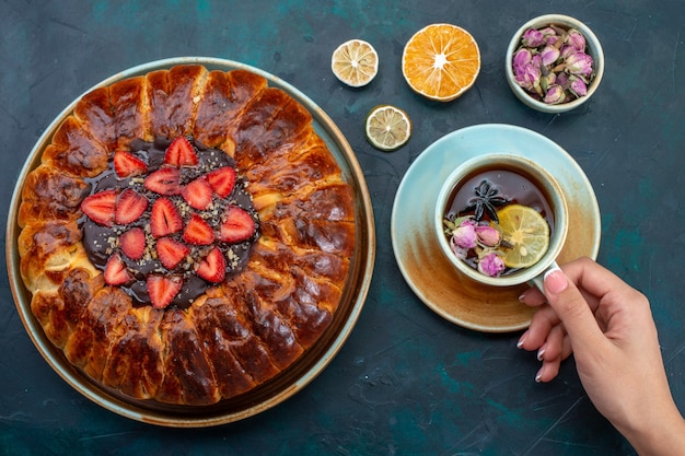 Вид сверху вкусного клубничного пирога с чашкой чая на темно-синей поверхности