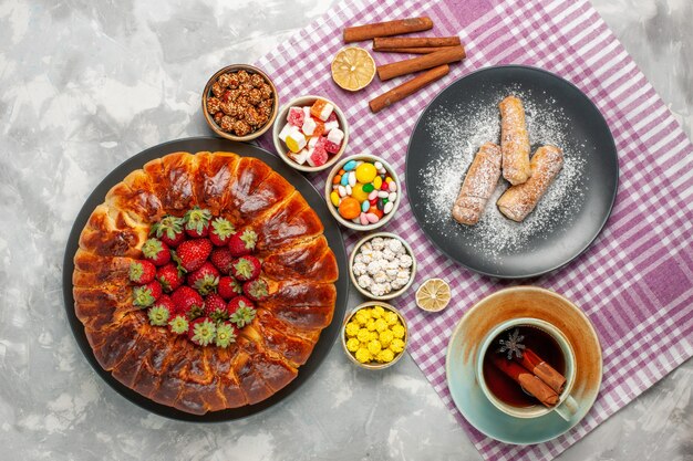 Top view of yummy strawberry pie with fresh red strawberries candies and cup of tea on white surface